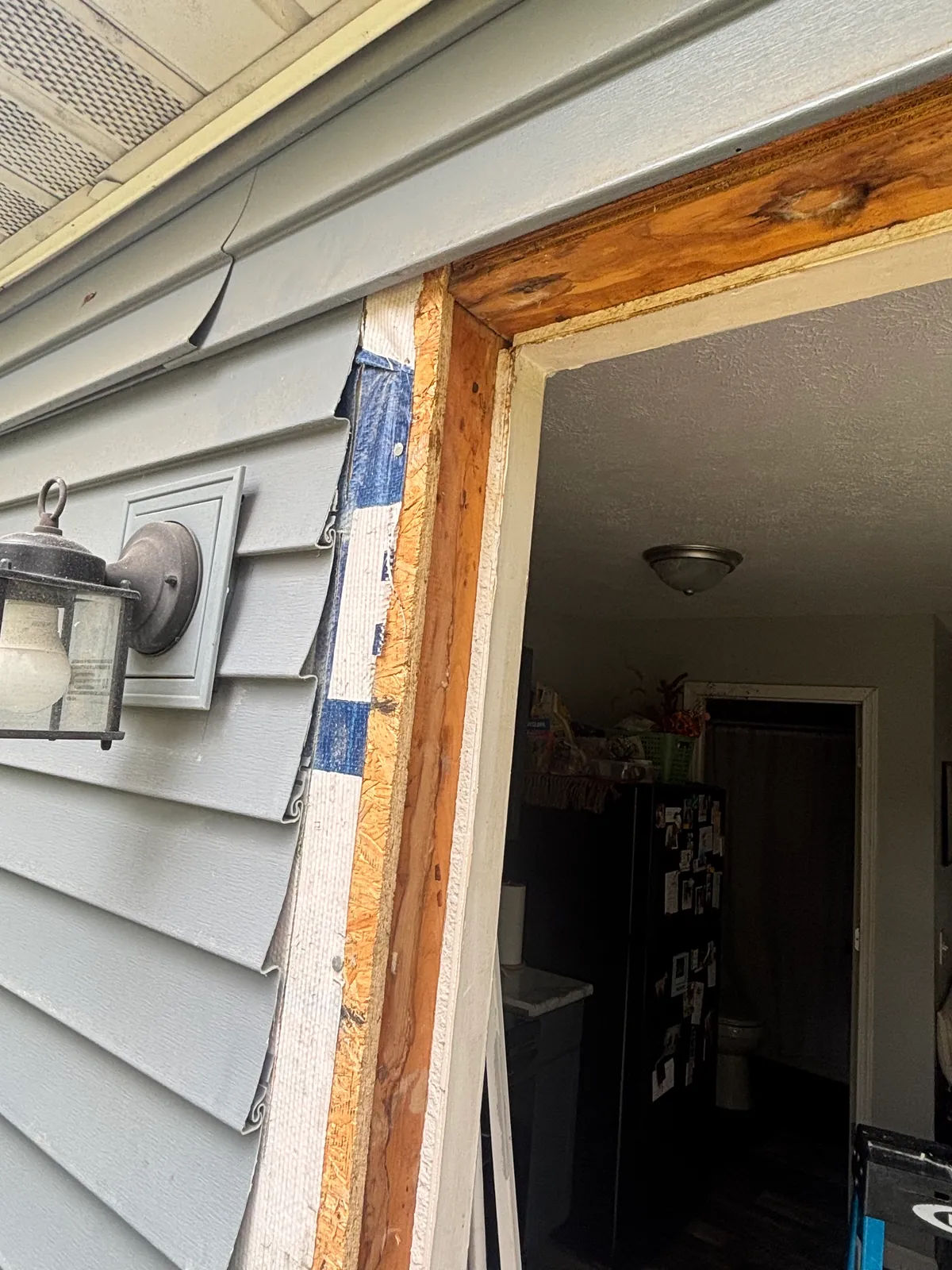 Door-sized opening framed in new lumber in the gray lap-sided wall of a house, with the OSB sheathing edge and housewrap seam tape exposed at the jamb and a wall lantern mounted alongside