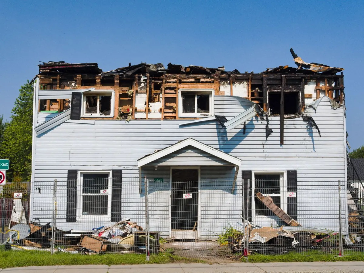 Two-story white-sided house with its upper floor and roof burned out, charred framing exposed and debris fenced off in the yard