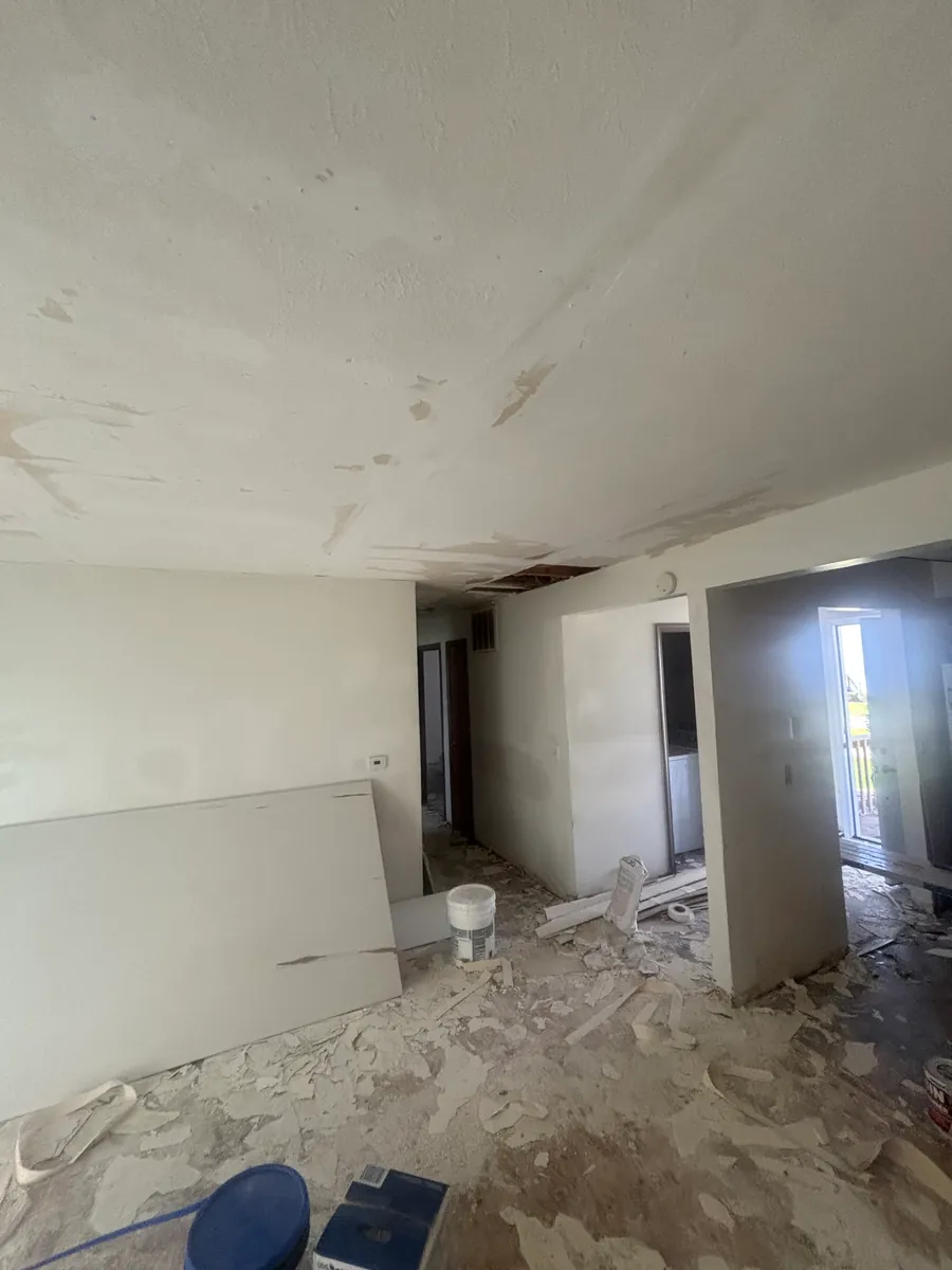 Emptied room mid-repair with a rectangular section of the ceiling cut away to the boards above, the surrounding ceiling skim-coated in joint compound, a sheet of drywall and a pail of compound standing by, and the removed material still covering the bare floor