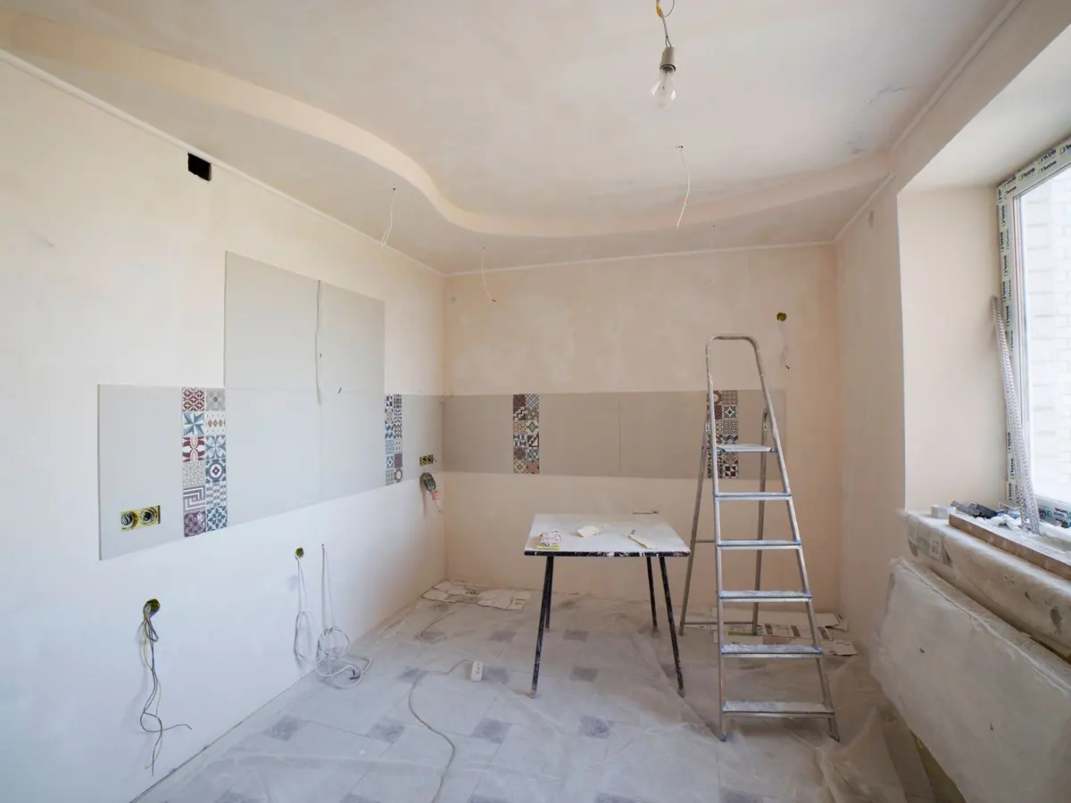 Empty room midway through an interior rebuild, with fresh drywall, exposed wiring boxes, a stepladder and covered floors