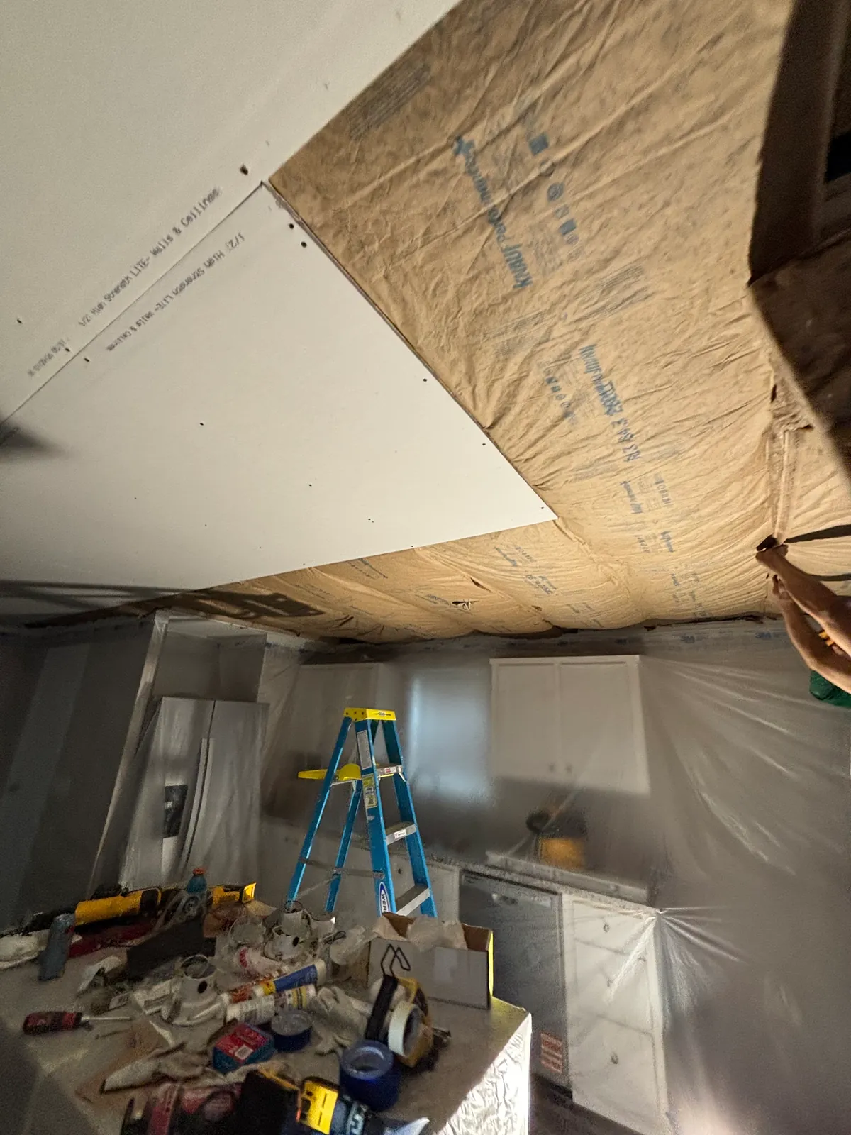 New drywall going up across a kitchen ceiling over exposed kraft-faced insulation, with a blue step ladder on the floor and the cabinets, refrigerator and island sealed behind plastic sheeting