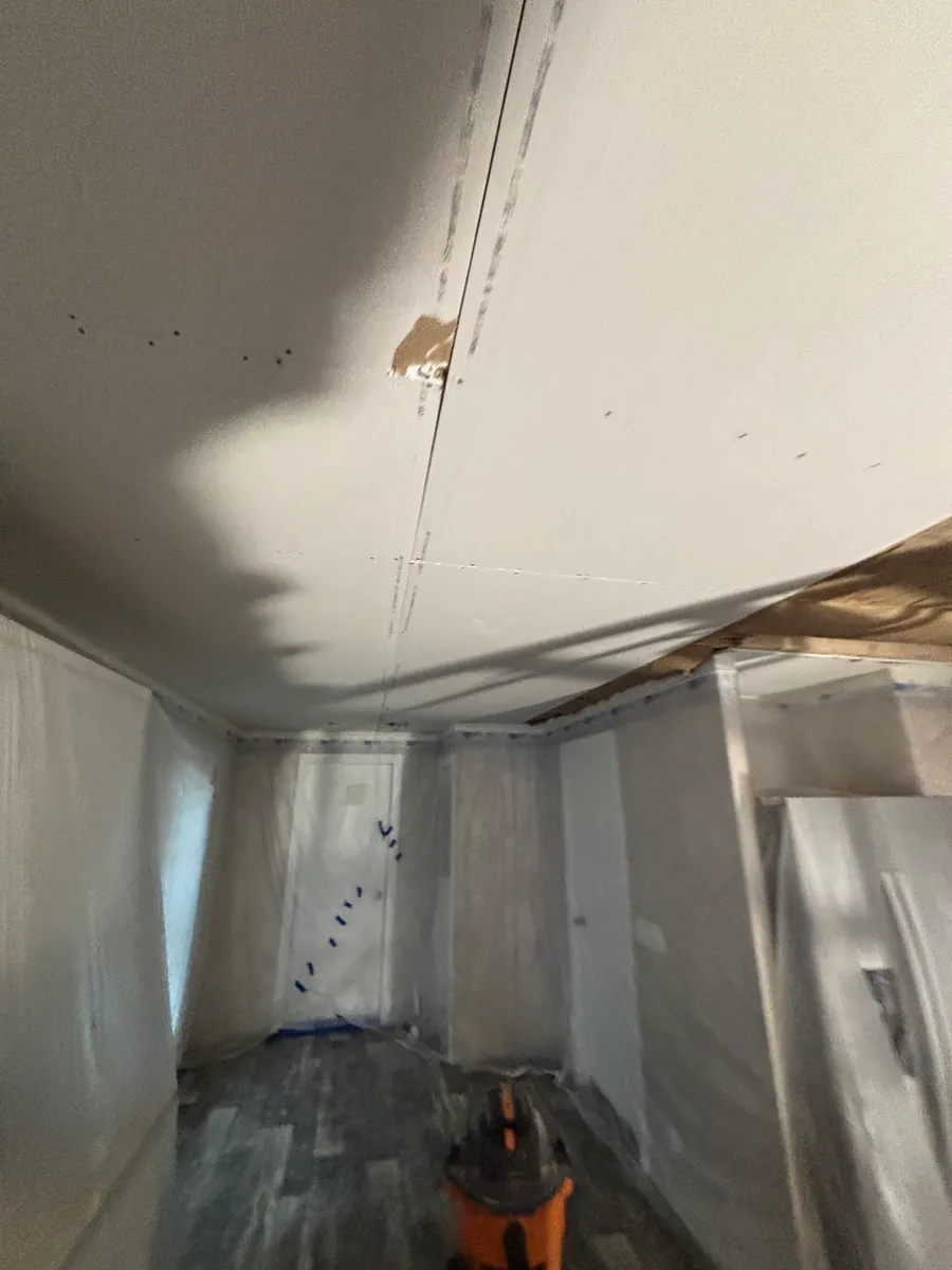Freshly hung and taped drywall ceiling in a room sheeted floor to ceiling in plastic containment, with an orange shop vacuum on the protected floor