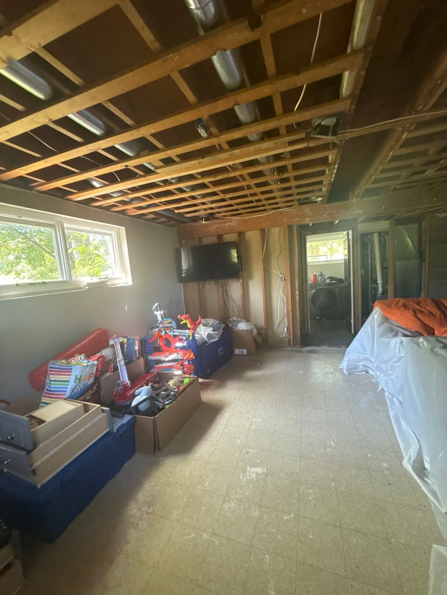 Room stripped for restoration with the ceiling drywall removed to expose joists, ductwork and wiring, one wall opened to the studs, and household boxes and totes stacked beside furniture covered in plastic sheeting