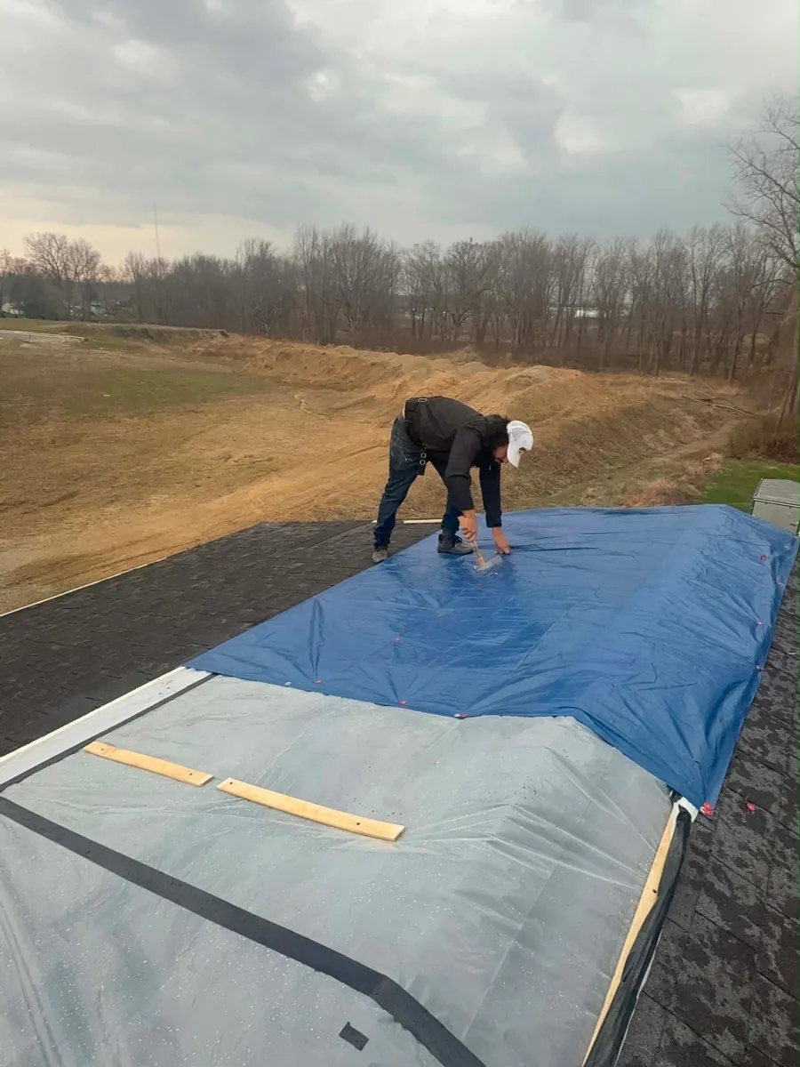 Lake Acer crew member in a white cap bent over a shingle roof driving a cap nail through the edge of a blue tarp, with clear plastic sheeting held down by wood battens on the slope below and bare winter trees beyond