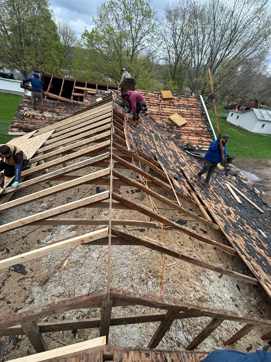 Four Lake Acer crew members working a roof tear-off, the near slope stripped back to bare rafters while old shingles and decking are cleared from the far slope beside the chimney