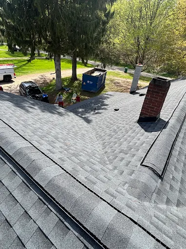 Finished gray asphalt shingle roof with a brick chimney, a job-site dumpster parked in the driveway below