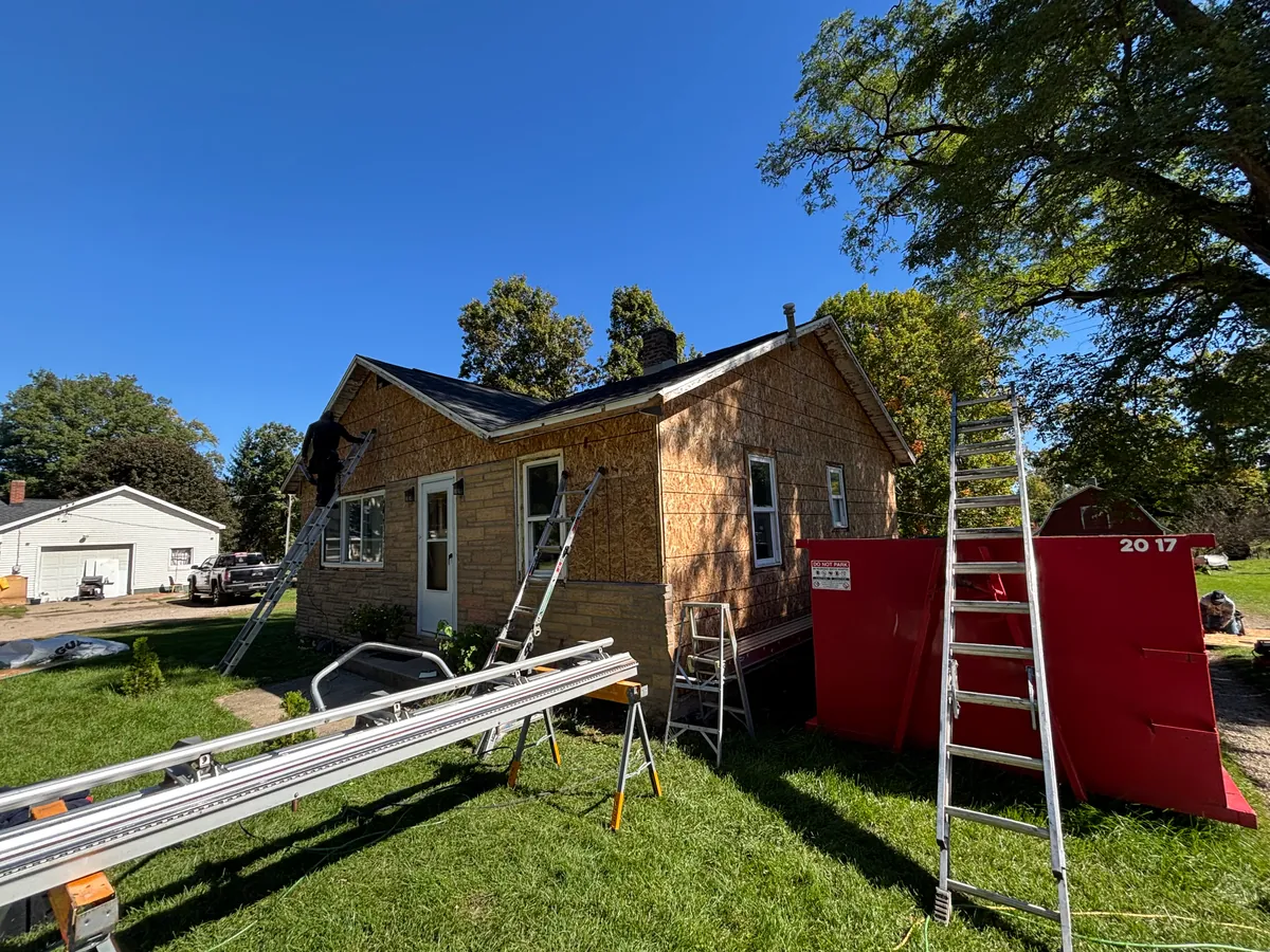 Single-story house stripped back to bare OSB sheathing below a finished charcoal shingle roof, with a crew member on an extension ladder at the gable end and a red roll-off dumpster in the yard